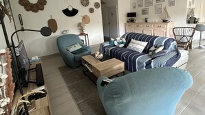 Un salon moderne avec un canapé, un fauteuil et une table basse. On y trouve également un téléviseur, une lampe et des œuvres d’art décoratives accrochées au mur.