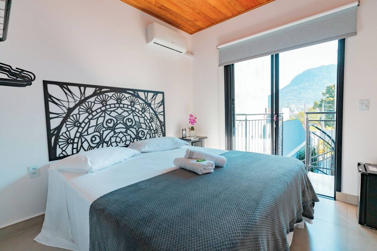 A modern bedroom with a large bed, a decorative headboard, a small bedside table with a plant, and a balcony with a view of the mountains.