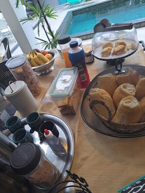 A breakfast setup with a fruit bowl, juice, cereal, and pastries on a table by a pool. - Hospedaria da Barra Balneário Camboriú  (Balneário Camboriú)