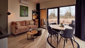 A modern living room with a brown sofa, a round wooden table, and black chairs. There is a large window with a view of trees and a fence. -  Hofparken De Bergvennen  (Lattrop)
