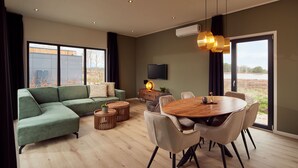 A modern living room with a green sectional sofa, a wooden dining table with chairs, and a large window offering a view of the outdoors. -  Hofparken De Bergvennen  (Lattrop)