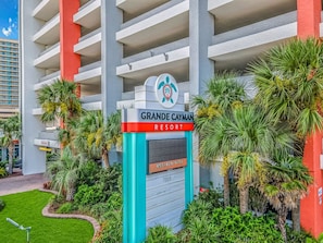 Entrada de un hotel con un letrero que dice Grande Cayman Resort, rodeado de palmeras y un césped cuidado.