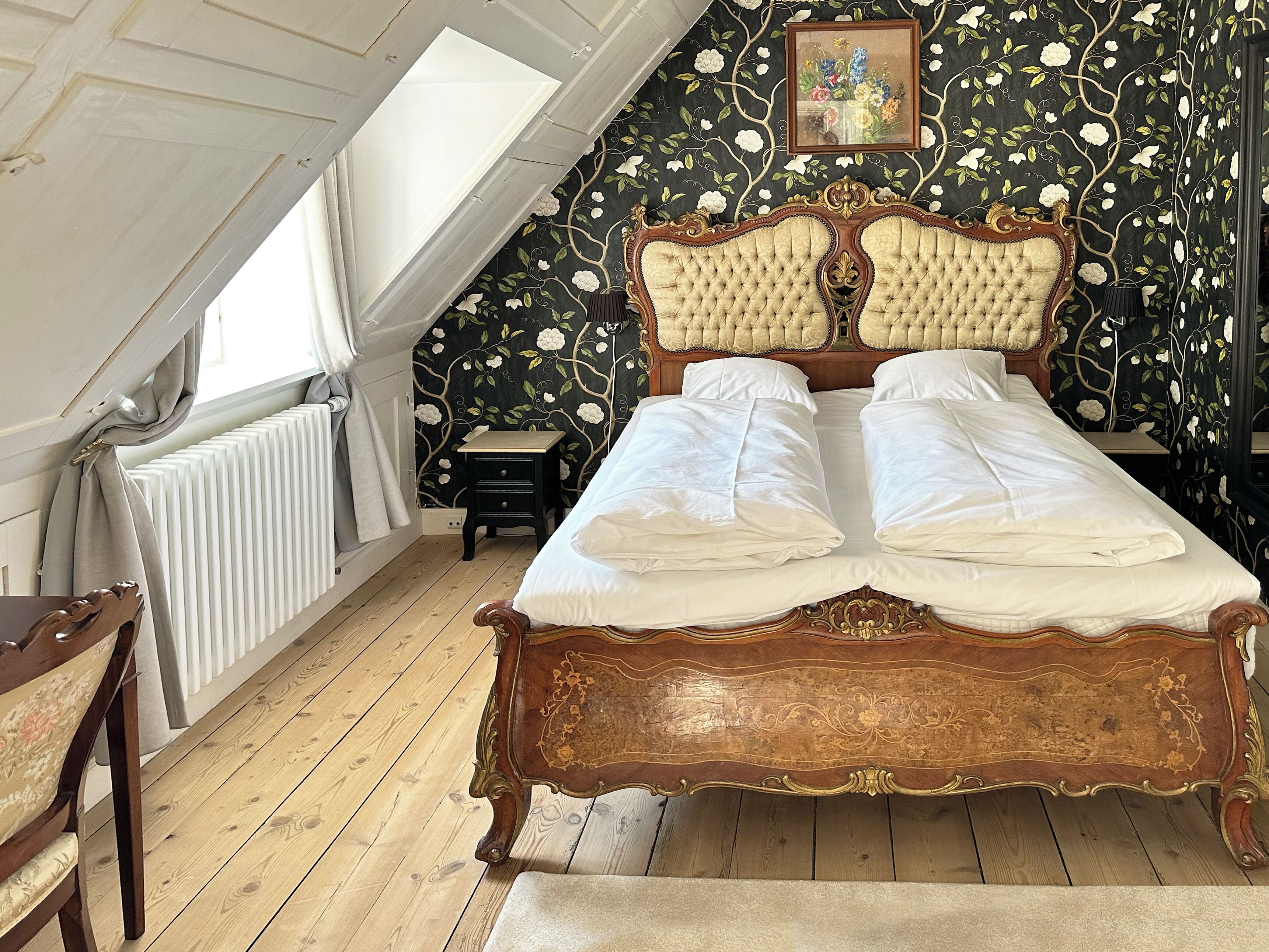 A bedroom with a wooden bed, a small bedside table, a chair, a radiator, and a window with curtains.