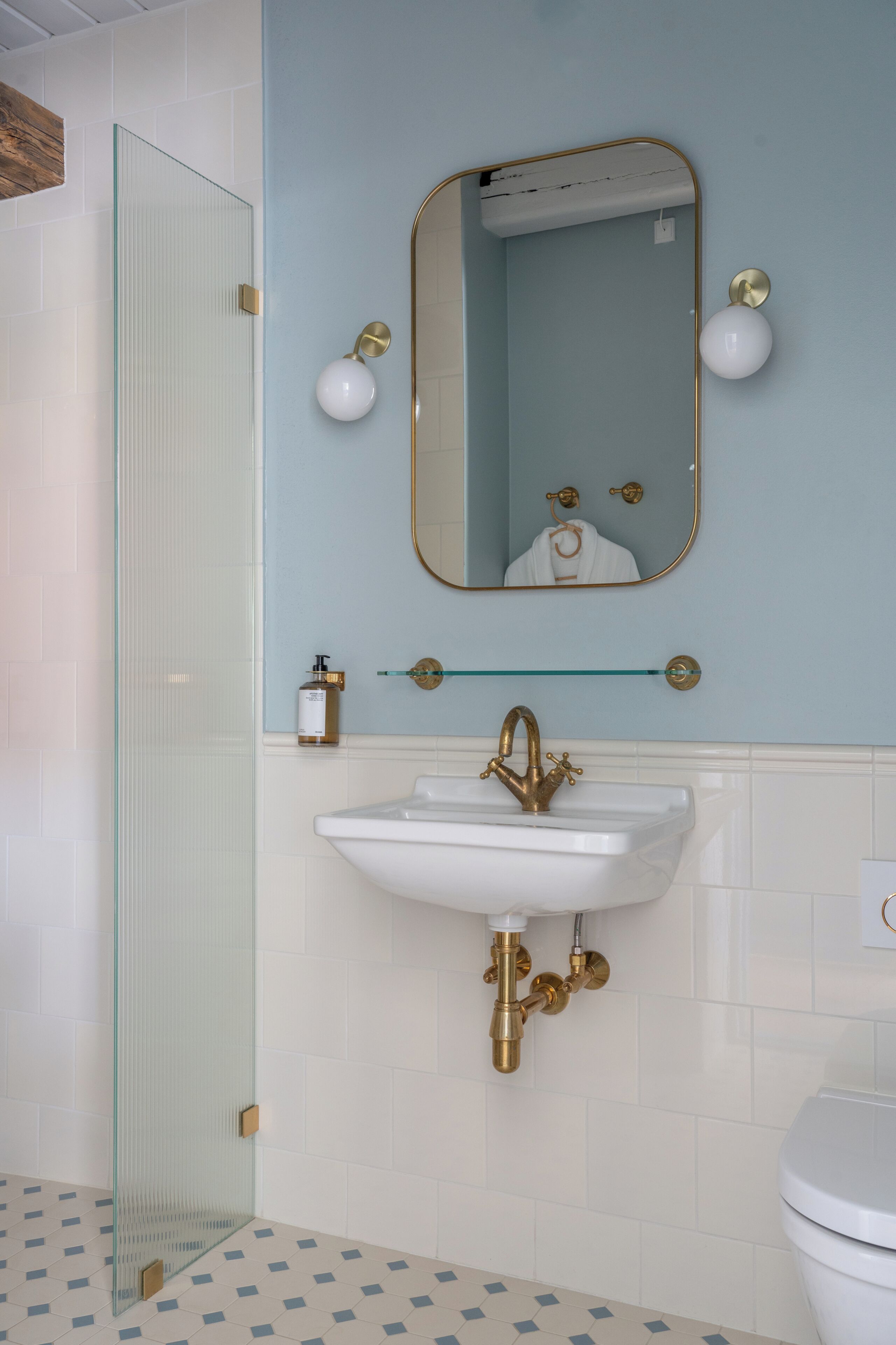 A bathroom with a glass shower enclosure, a white sink with a gold faucet, a mirror with gold-framed surround, and a toilet.