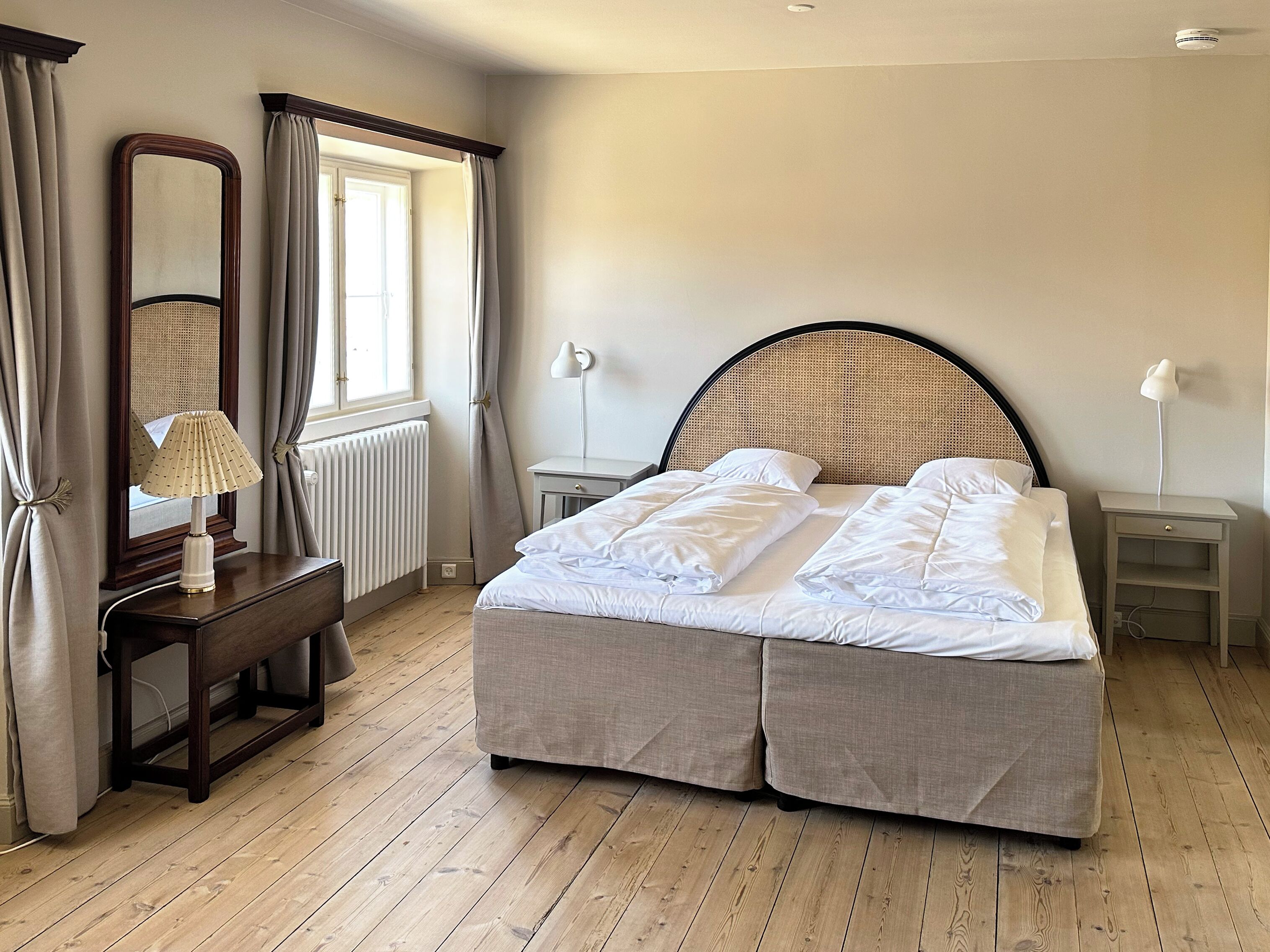 Un dormitorio con cama, mesita de noche, lámpara, espejo y ventana con cortinas.