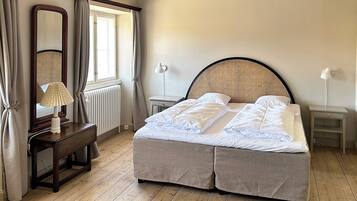 Un dormitorio con cama, mesita de noche, lámpara, espejo y ventana con cortinas.