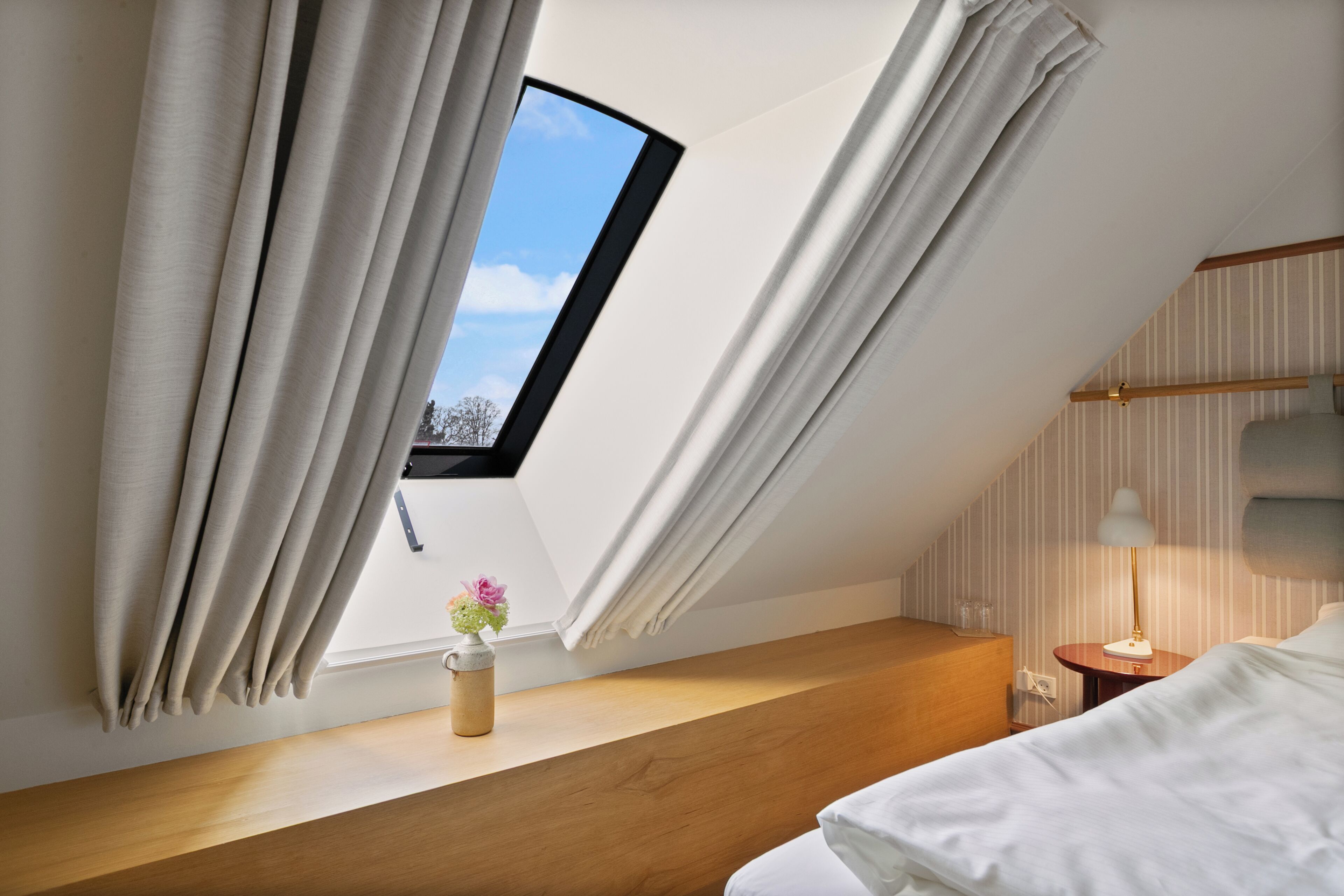 A bedroom with a skylight, a bedside table with a lamp, and a window with curtains.