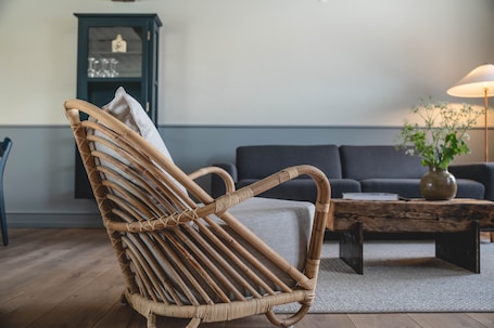 A wicker chair with a cushion, a dark sofa, a wooden coffee table, and a lamp.. Broløkke Herregård