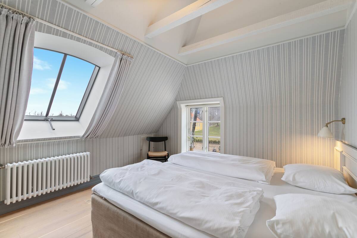 A bedroom with a sloped ceiling, a large window, a radiator, and a bed with white bedding.