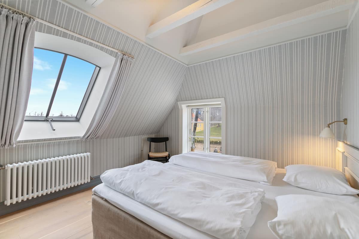 Une chambre avec un plafond en pente, une grande fenĂȘtre, un radiateur et un lit avec une literie blanche.