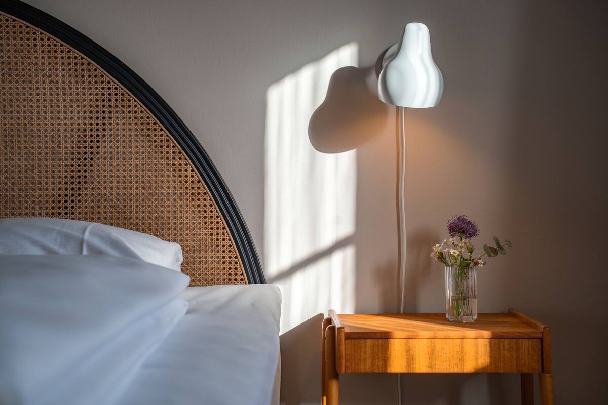 A neatly made bed with white linens, a wooden bedside table with a vase of flowers, and a wall-mounted lamp.