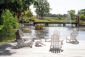 Una terraza de madera con sillas Adirondack, un pequeño edificio amarillo y un puente sobre un cuerpo de agua tranquilo.