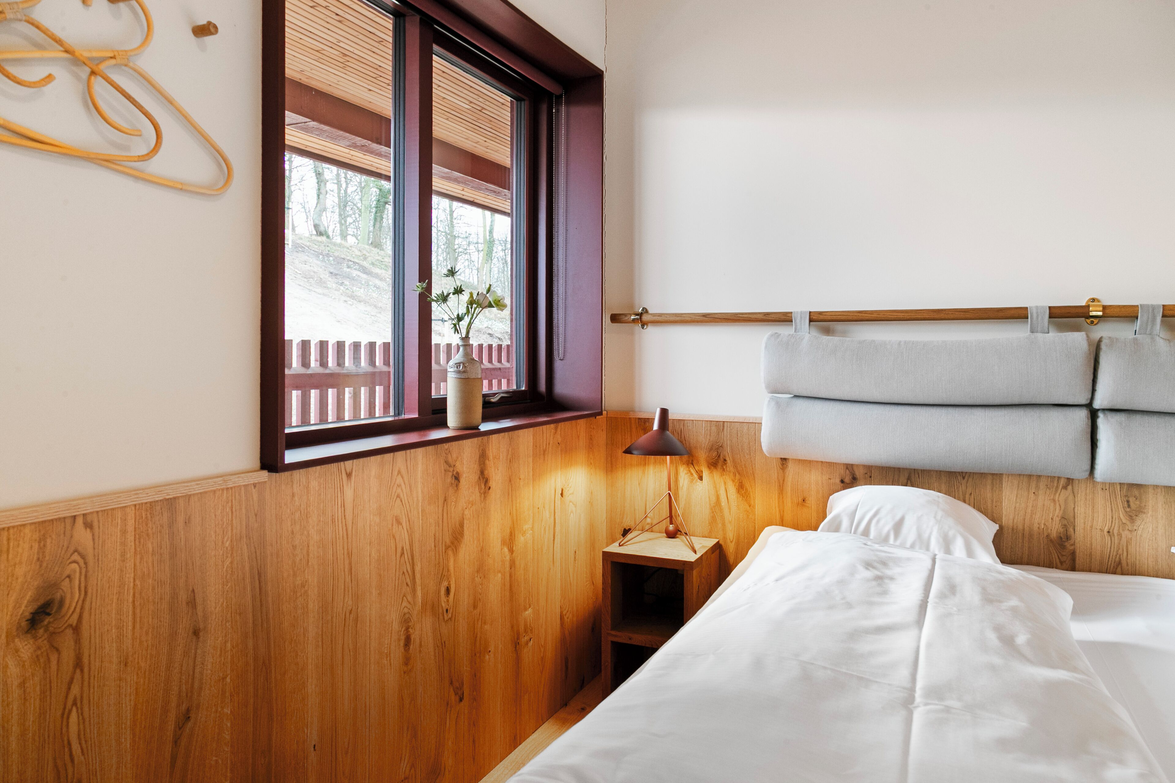 Un dormitorio con cabecera de madera, una mesita de noche con lámpara, una ventana con persianas y vistas a una valla y árboles.