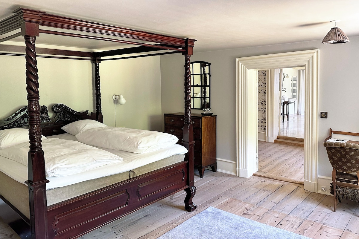 Una cama con dosel de madera y una colcha blanca en una habitación con suelo de madera y una puerta blanca que da a otra estancia.