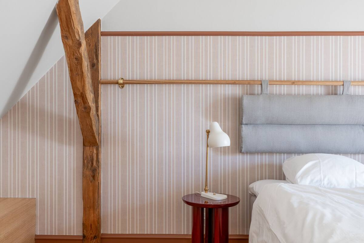 A bedroom with a sloped ceiling, a bed, a bedside table with a lamp, and striped wallpaper.