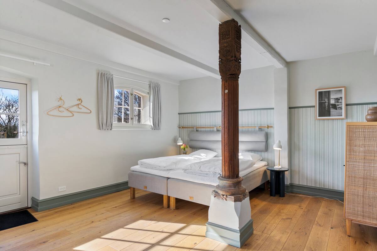A bedroom with a bed, a wooden pillar, a window with curtains, a bedside table, and a lamp.