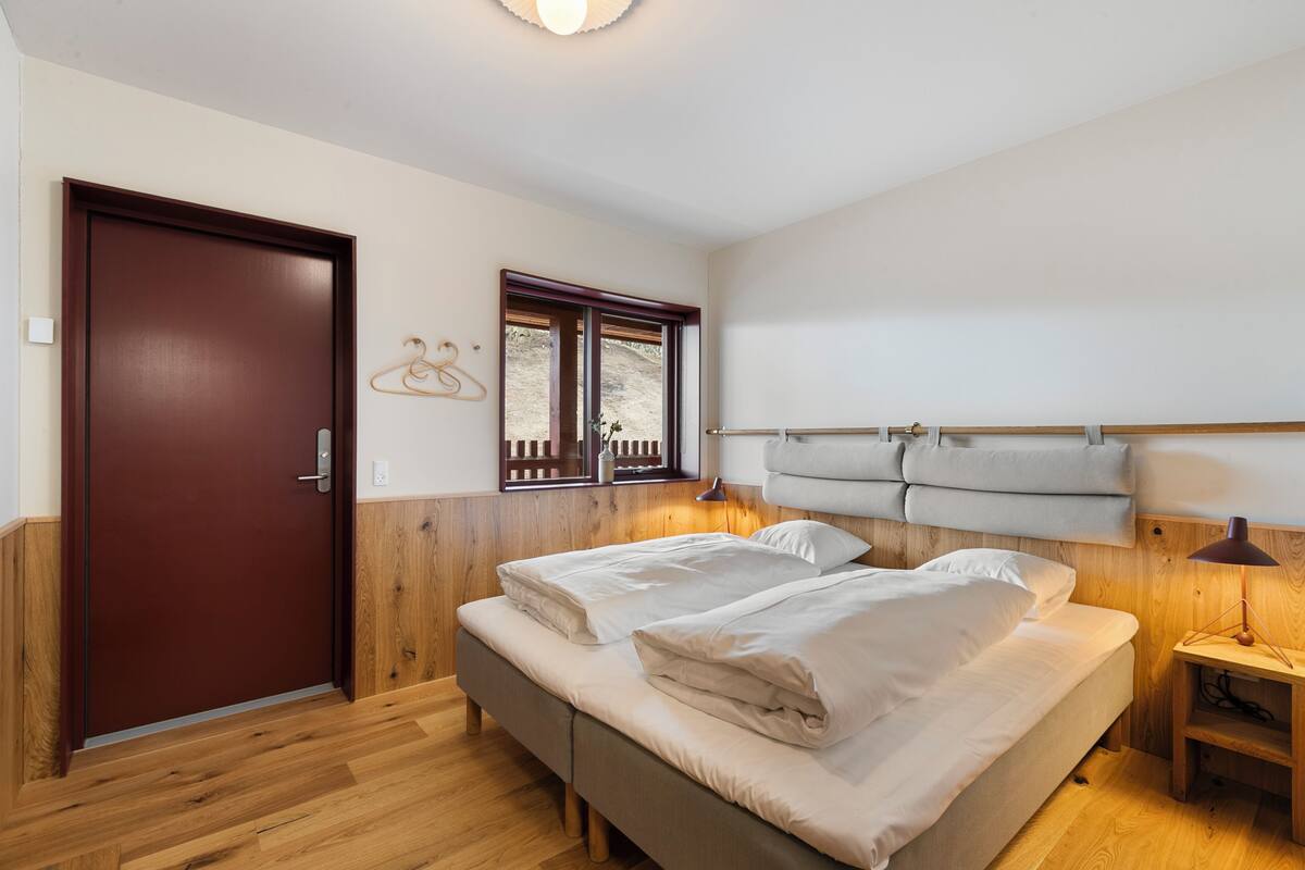 A bedroom with a wooden door, a window, a bed with pillows, a bedside table, and a hanging light fixture.