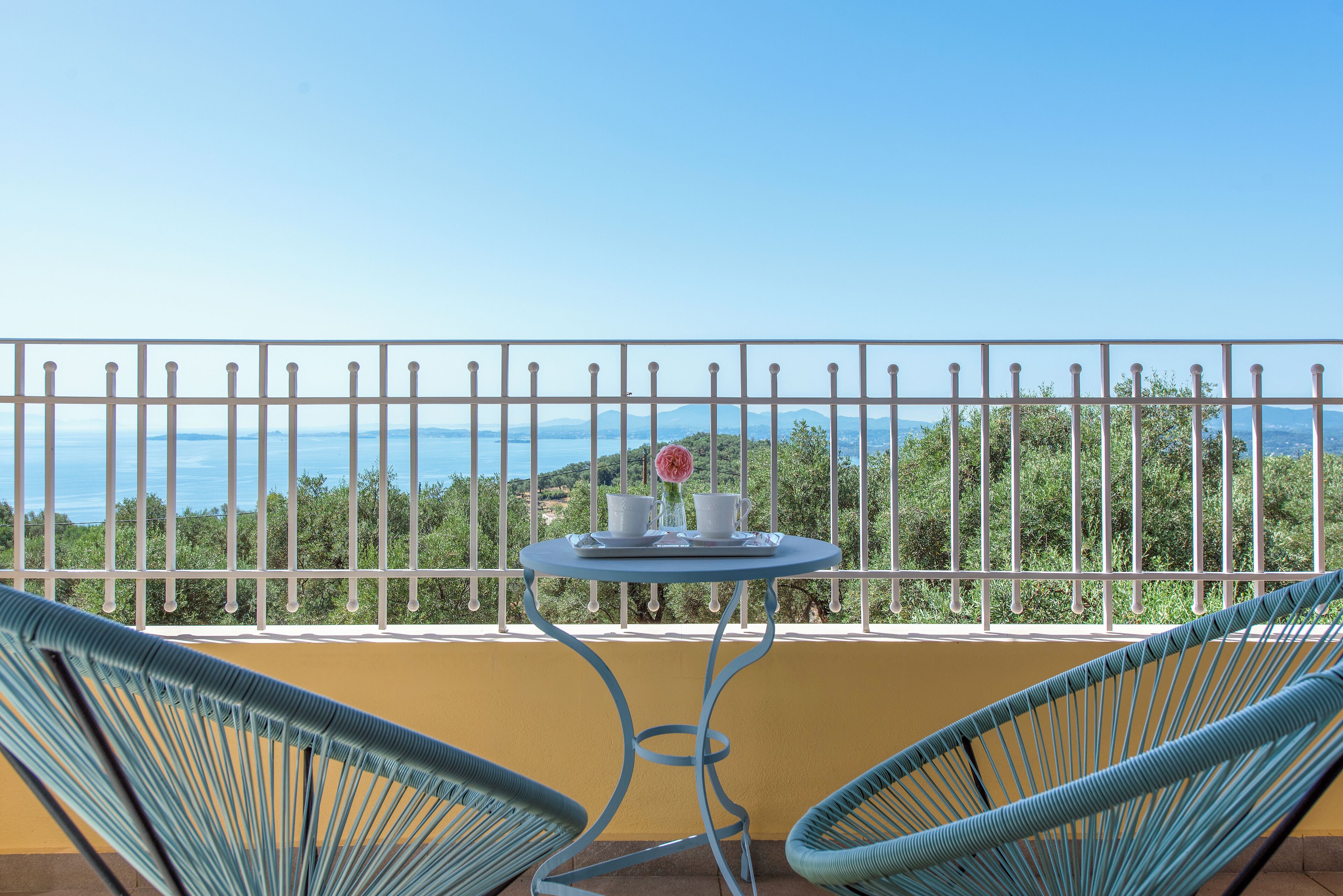 A balcony with a table and chairs, overlooking a scenic view of hills and the sea.
