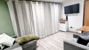 Ett modernt vardagsrum med grå soffor, ett trägolv, en platt-TV som är monterad på väggen och en liten köksdel med ett träbord och skåp.