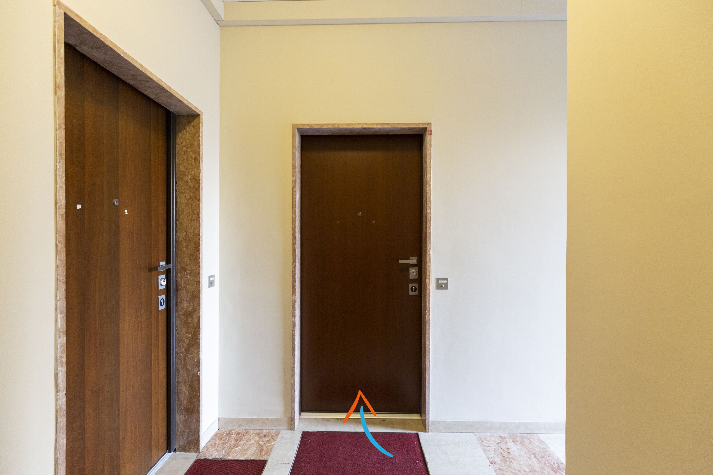 A hallway with two wooden doors, a marble floor, and a red doormat.