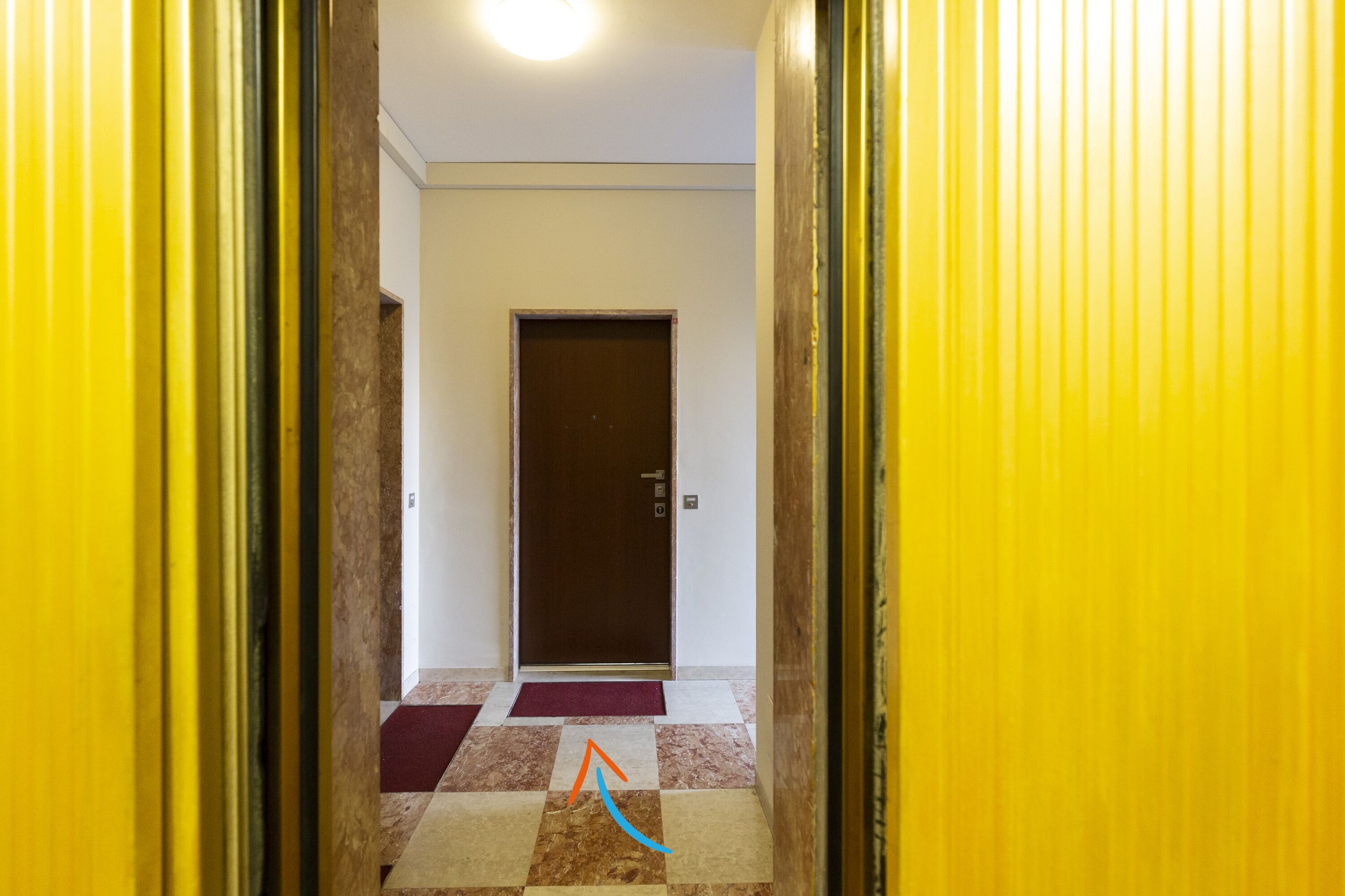 Un couloir avec un sol à carreaux, une porte marron au fond et des murs jaunes.