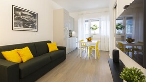 Ein modernes Wohnzimmer mit einem schwarzen Sofa, gelben Kissen, einem Essbereich mit einem weißen Tisch und gelben Stühlen sowie einer Küche im Hintergrund.