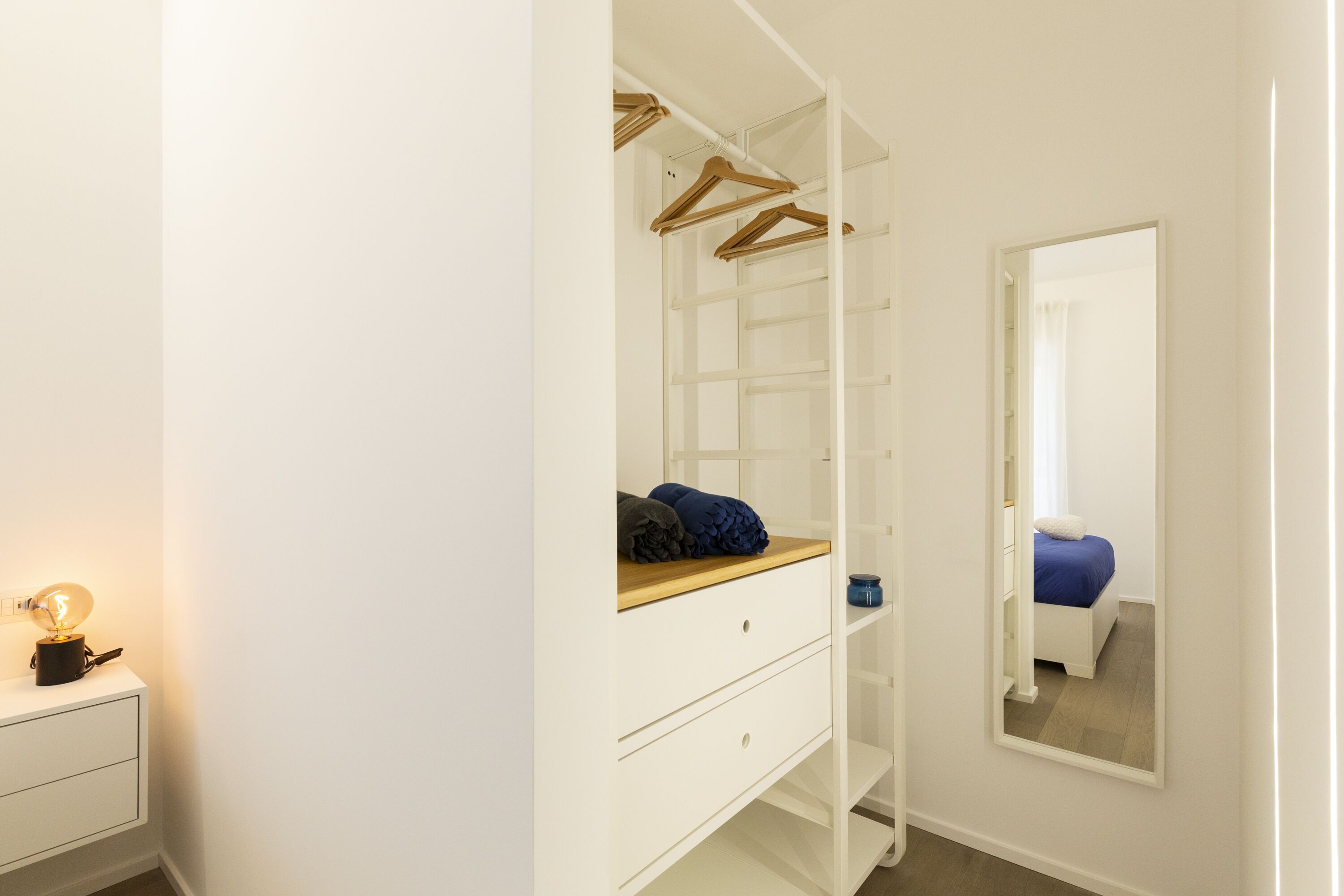 Un dressing accessible sans porte, doté d’étagères ouvertes, d’un miroir et d’une table de chevet avec une lampe.
