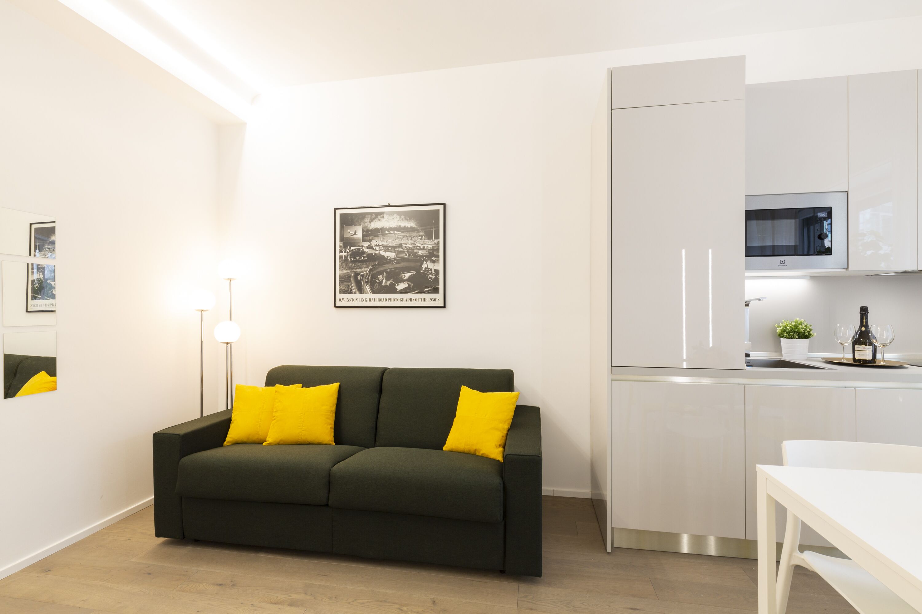 A modern living room with a dark green sofa and yellow cushions, a white dining table, and a kitchen area with white cabinets and a microwave.