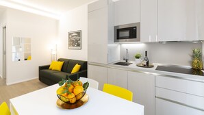 Eine moderne Küche mit weißen Schränken, einer Mikrowelle und einem Spülbecken. Ein Esstisch mit Obstschale und Stühlen steht daneben. Im Hintergrund ist eine gelb gepolsterte Couch zu sehen.