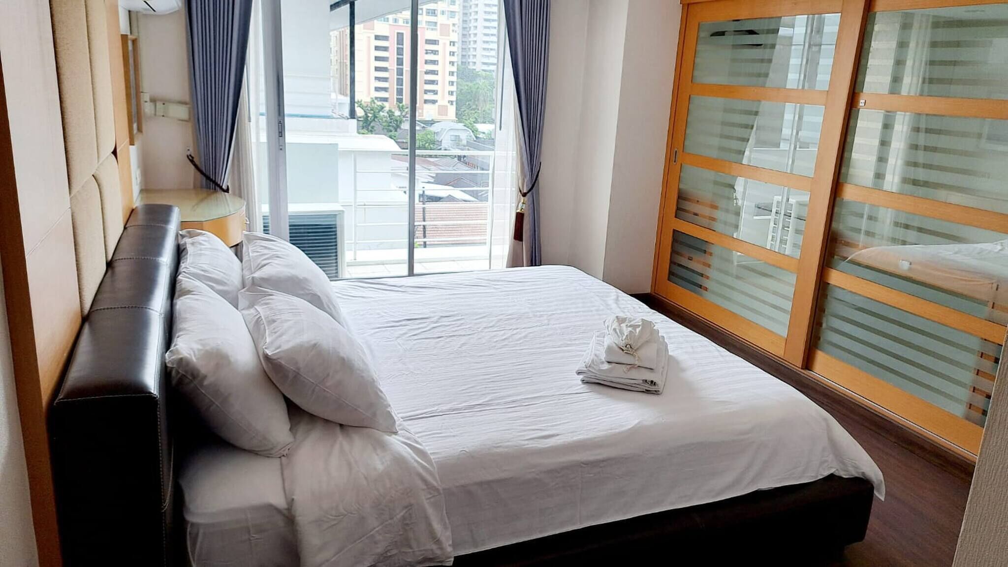 A hotel room with a bed, a headboard, a window with curtains, and a view of buildings outside.