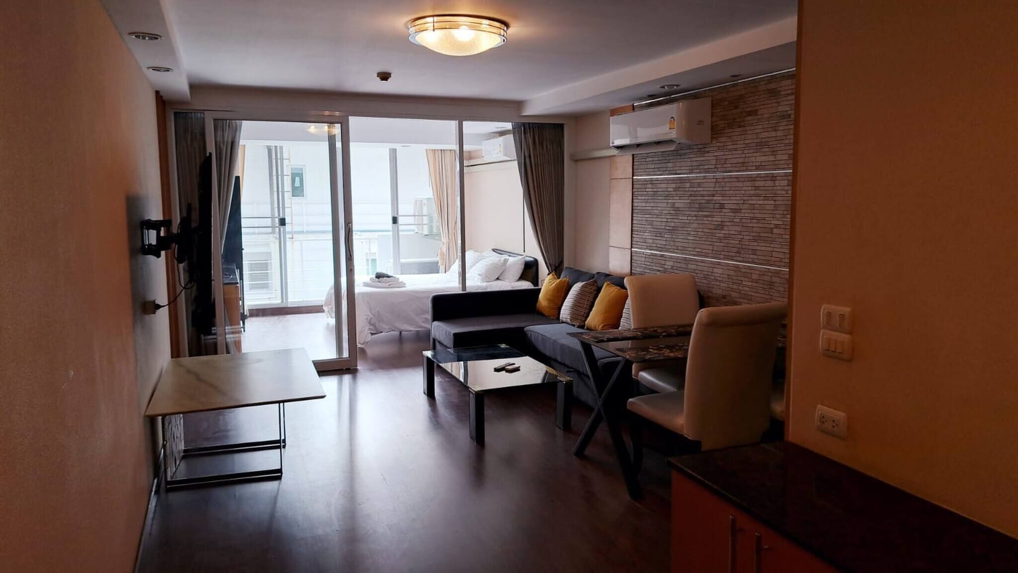 A modern hotel room with a sofa, armchair, and a small table. There is a balcony with a bed and a view of the city.