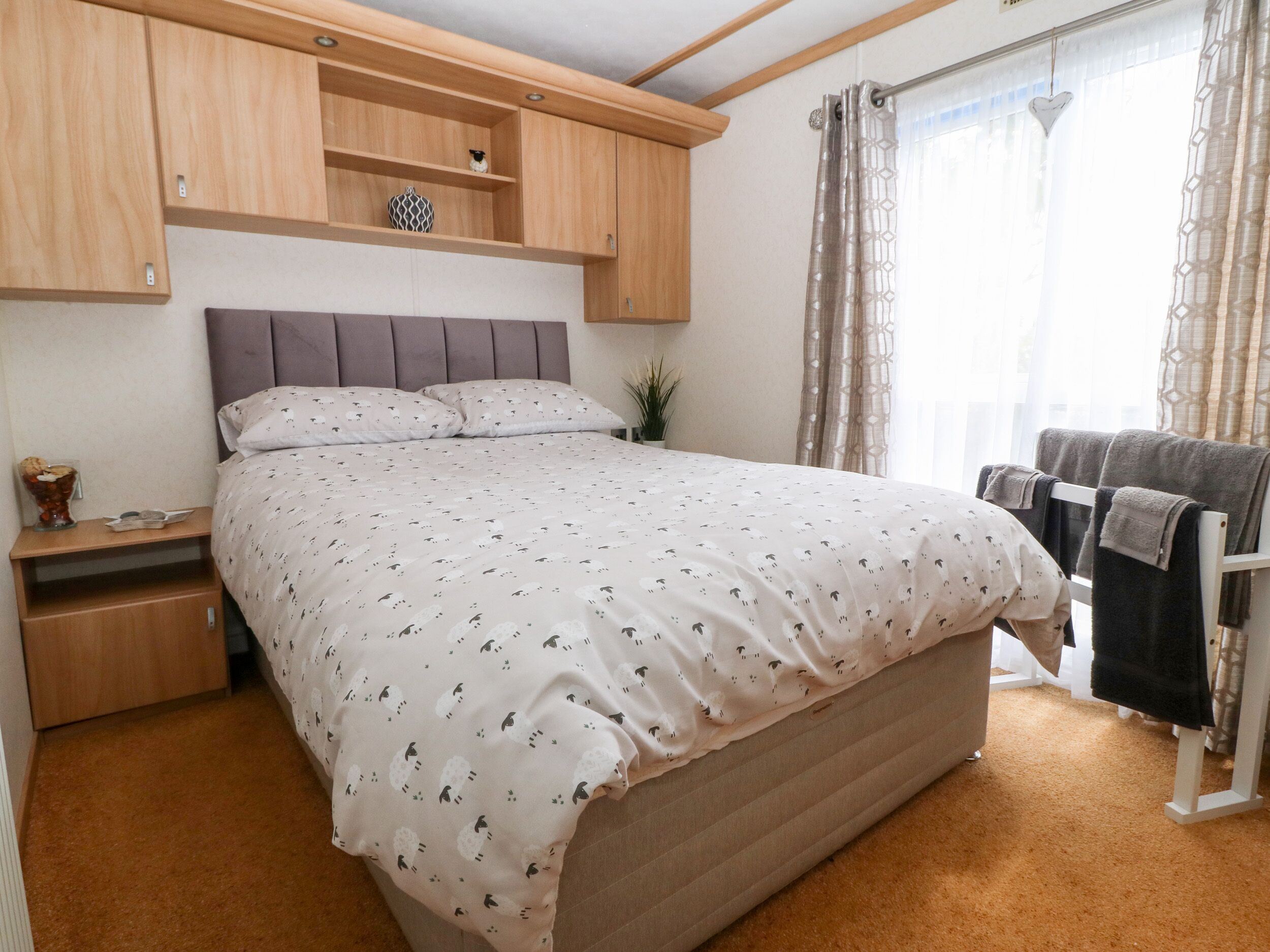 A bedroom with a bed, a nightstand, a shelf with a vase, and a window with curtains.