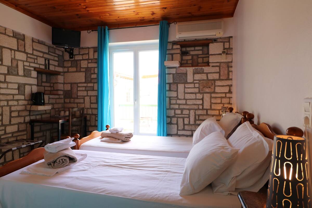 Une pièce avec un mur en pierre, un lit recouvert de draps blancs, un plafond en bois et une fenêtre avec des rideaux.