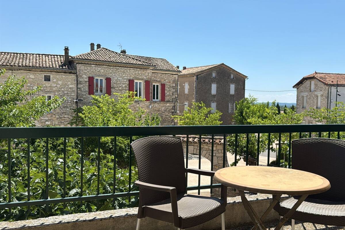 Un balcon agrémenté de chaises en osier et d’une petite table ronde, d’où l’on peut admirer des bâtiments en pierre aux volets rouges et un ciel d’un bleu limpide.