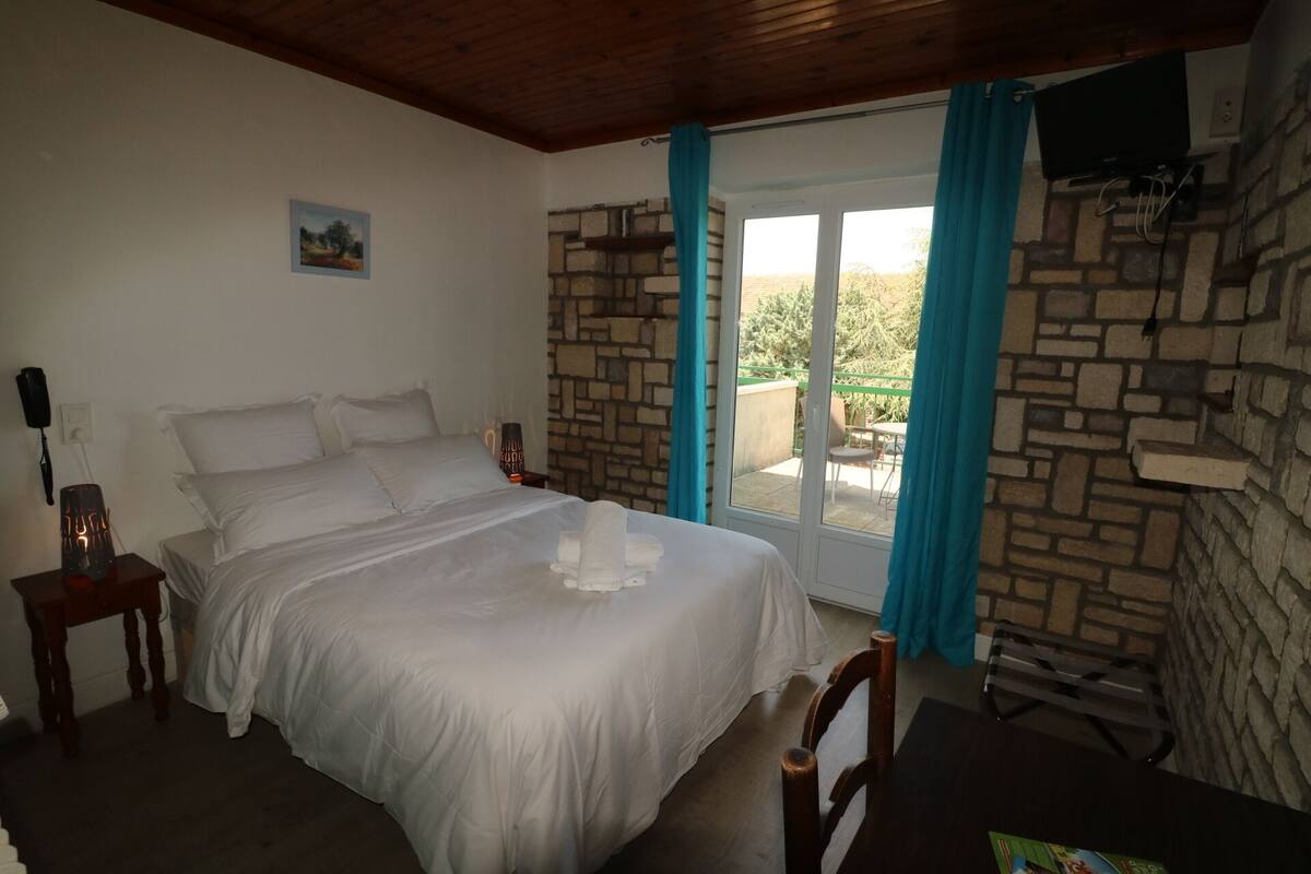 Une chambre à coucher avec un grand lit, une tête de lit en bois, une table de chevet, une lampe et une vue sur l’extérieur grâce à une porte vitrée.