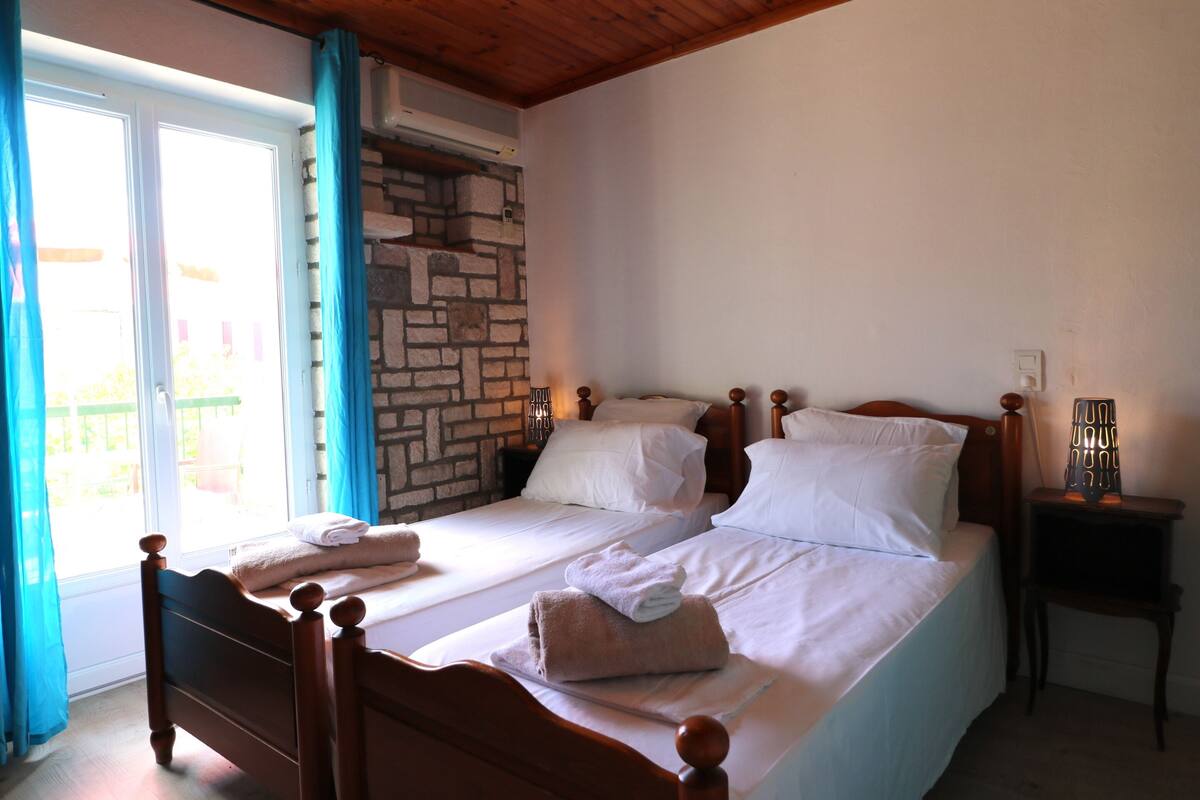 Une chambre à coucher avec deux lits en bois, un mur en pierre, une fenêtre avec des rideaux et une table de chevet avec une lampe.