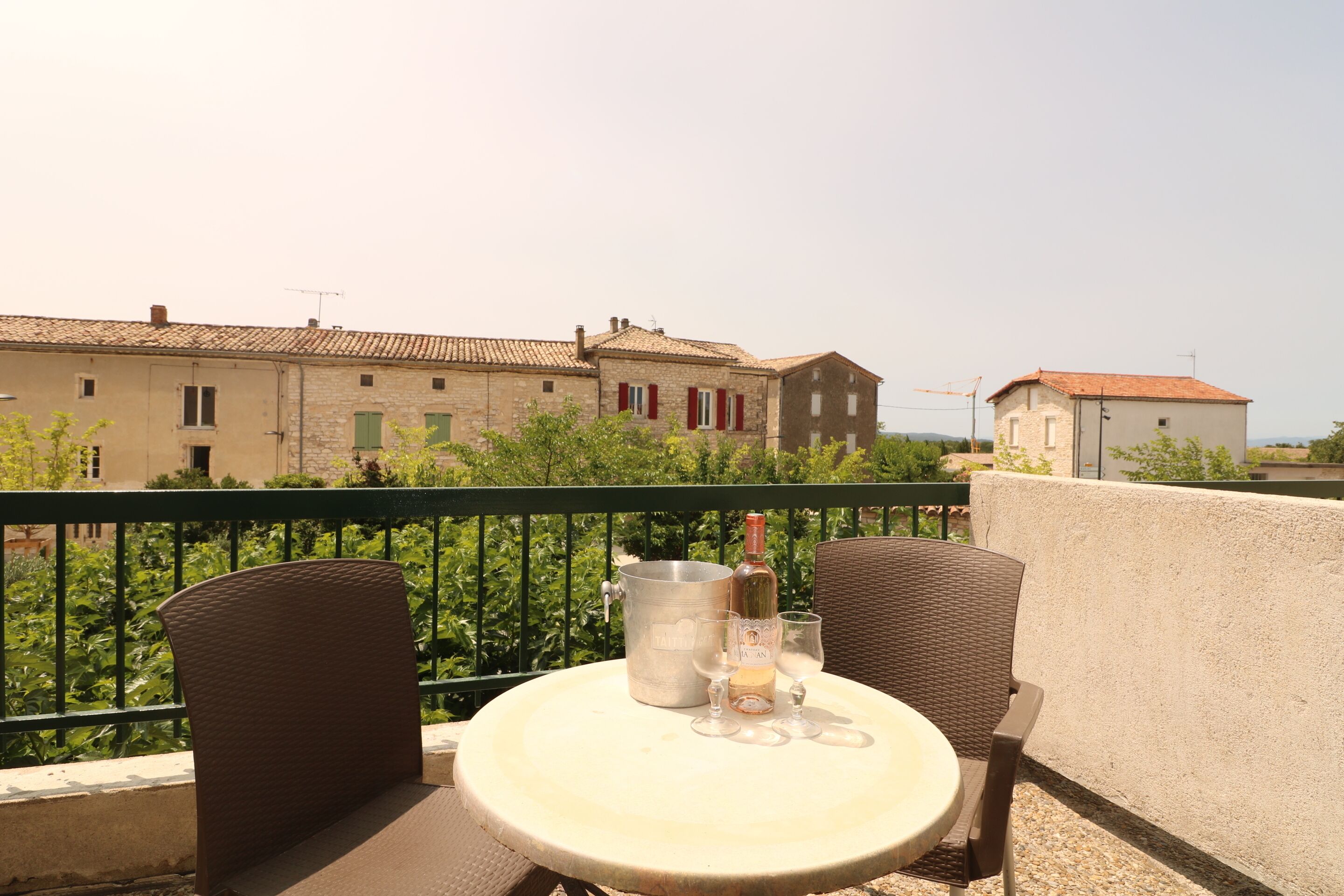 A balcony with two wicker chairs, a round table, a wine bottle, and glasses. In the background, there are buildings with red shutters and a clear sky.