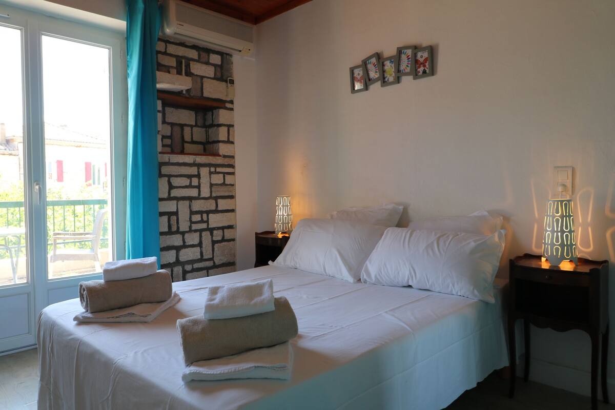 Une chambre à coucher avec un lit, une table de chevet, une lampe fixée au mur et une vue sur l’extérieur, visible à travers une porte vitrée.