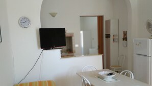 Una habitación compacta con una mesa de comedor, sillas, un televisor montado en la pared, un refrigerador y un reloj.