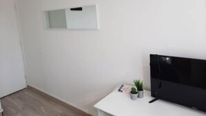 A minimalist room with a white wall, a mounted TV, a small white desk, and two potted plants. - Appartement T4 Rouen (Rouen)