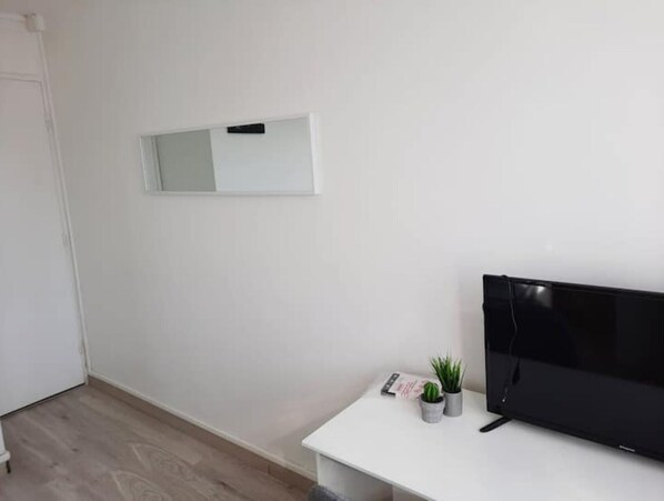 Une pièce au décor minimaliste, avec un mur blanc, un téléviseur fixé au mur, un petit bureau blanc et deux plantes en pot.
