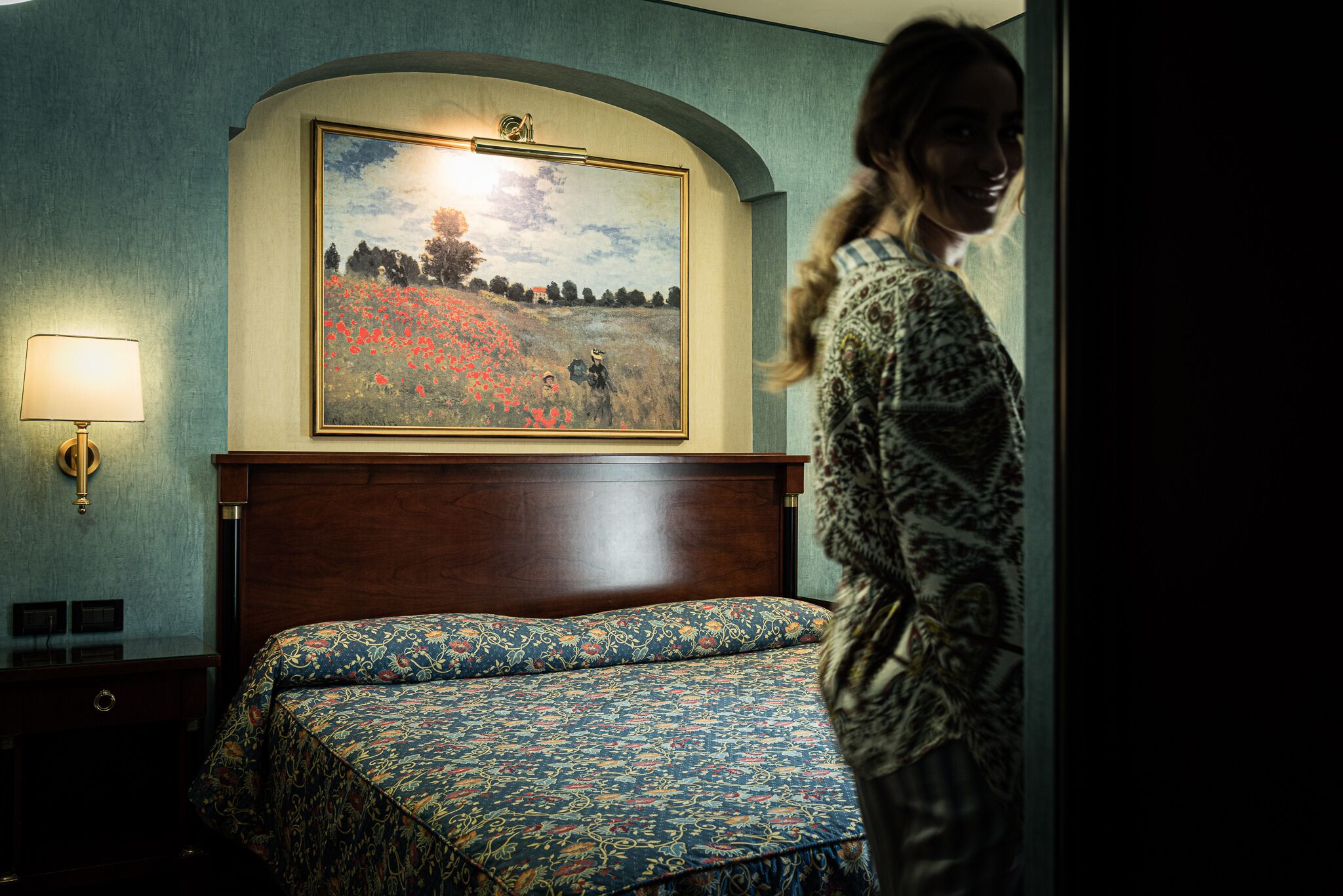 A hotel room with a bed, a painting on the wall, and a lamp.