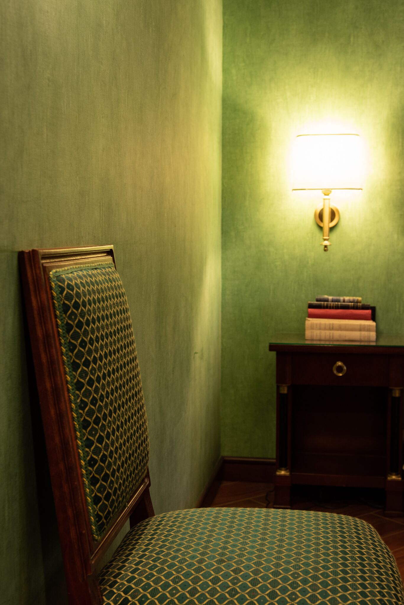Una habitación con pared verde, una silla de madera con cojín estampado, una pequeña mesita de madera con libros y una lámpara fijada a la pared.