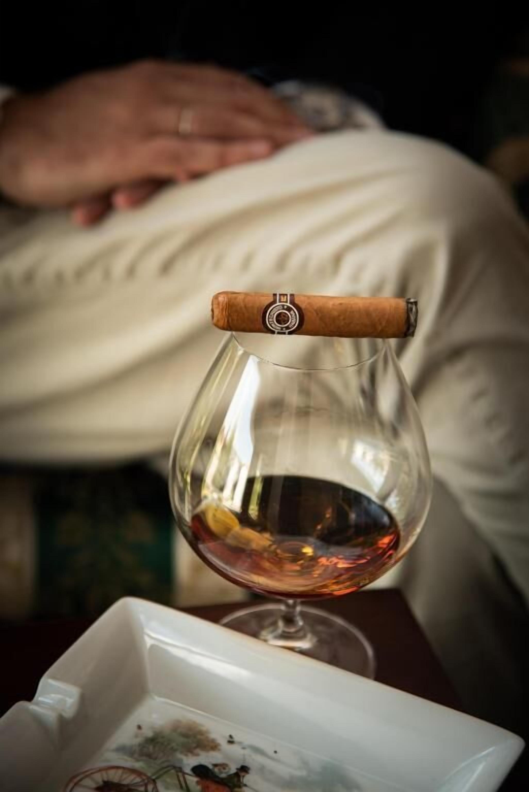 A glass of amber liquid with a cigar on the rim, placed on a table with a white tray containing a plate with remnants of food.