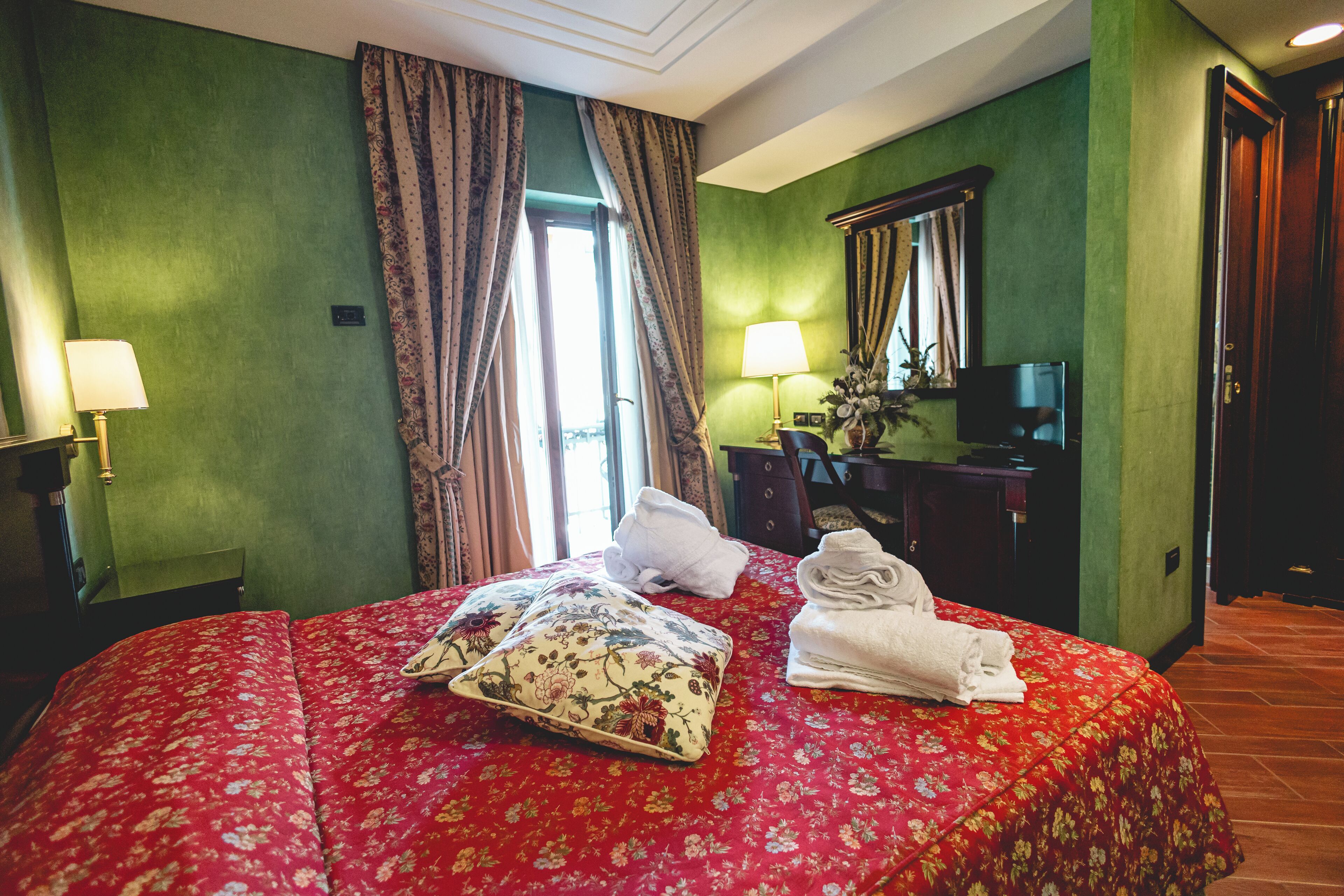 A hotel room with a floral bedspread, a desk with a lamp, a television, and a mirror on the wall.