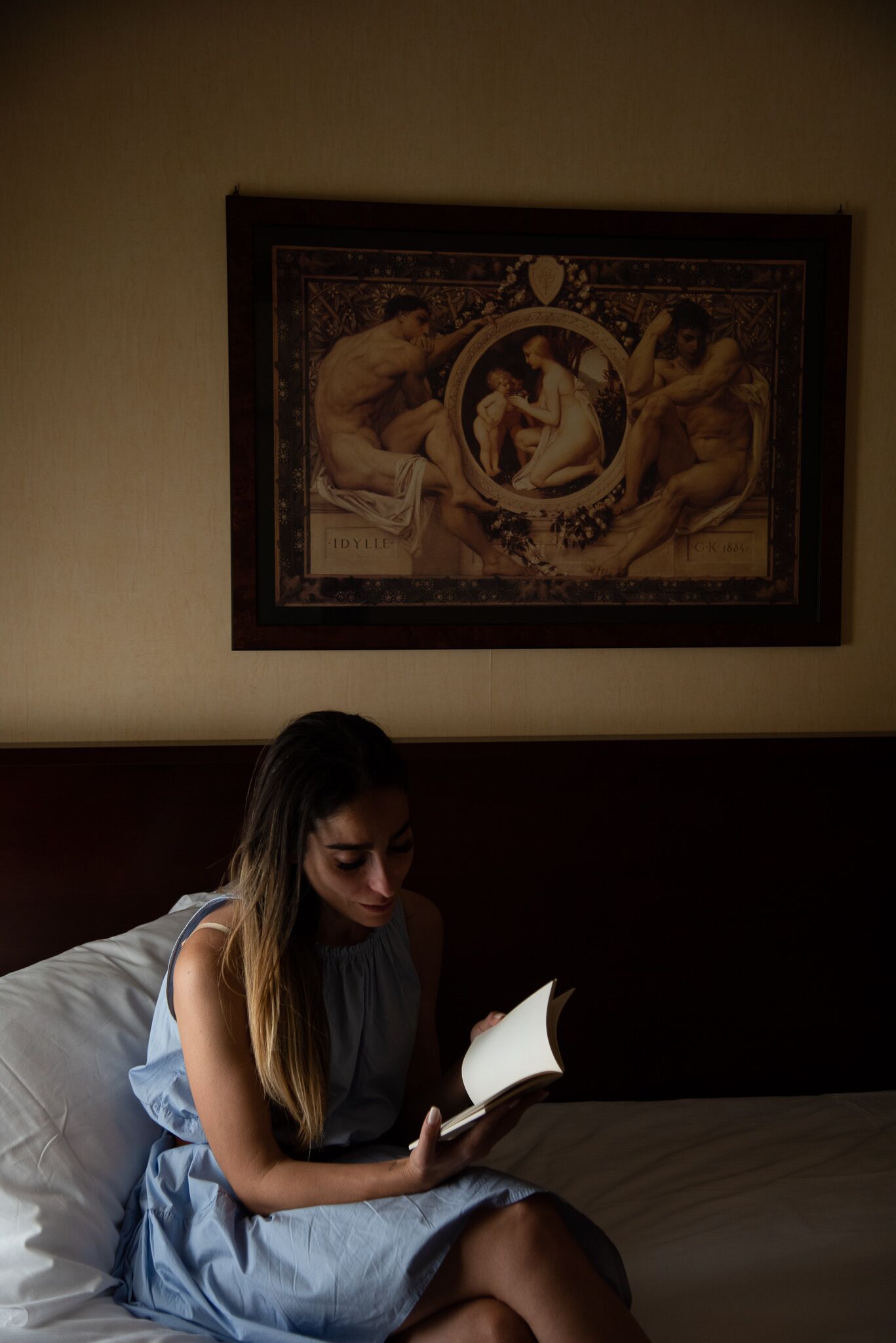 Una habitación con una cama, una persona leyendo un libro y una obra de arte enmarcada en la pared.