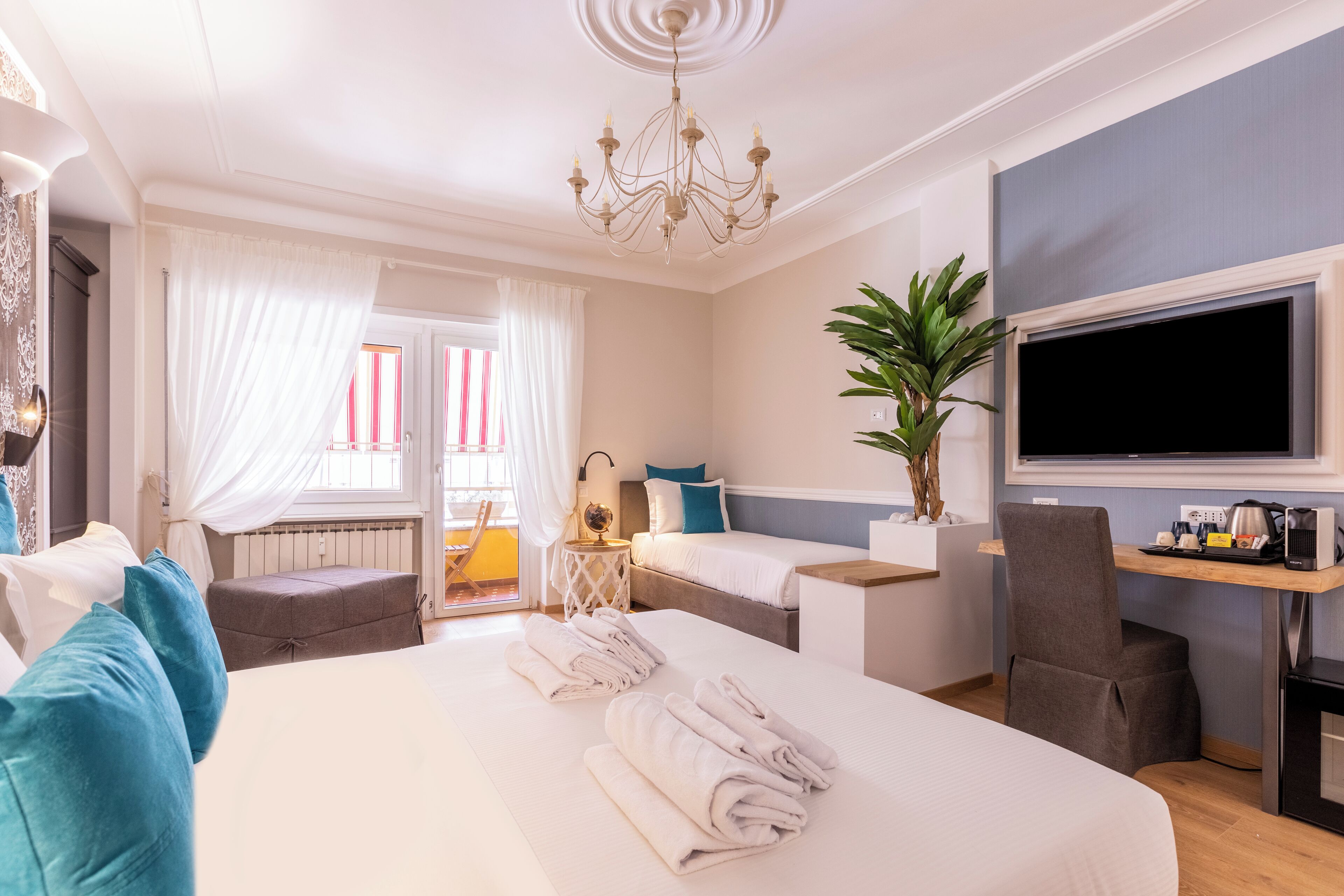 A hotel room with a large bed, a desk with a flat-screen TV, a chair, a small table with a globe, and a potted plant.