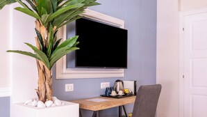 A room with a wooden desk, a grey chair, a large plant, a flat-screen TV, and a white door. - Trastevere Paper Suites (Rome)