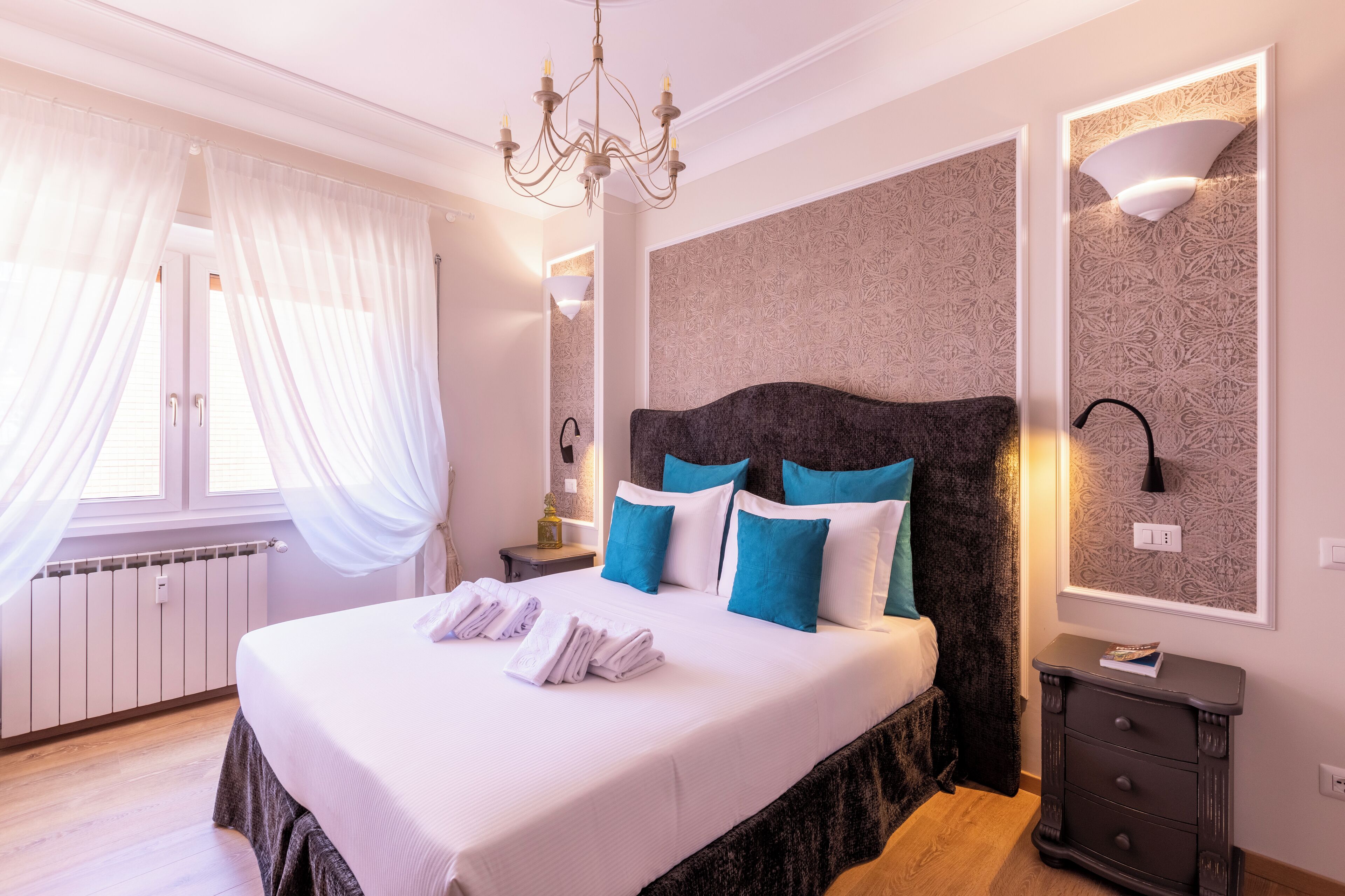 A bedroom with a large bed, a chandelier, a bedside table, and a wall-mounted light fixture.