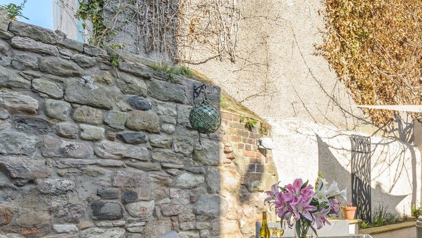 Un mur de pierres, des meubles en osier et une table avec une bouteille et des fleurs.